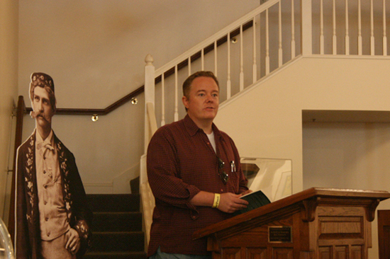 Paul Miles Schneider reads from his novel “Silver Shoes” at the historic Columbian Theatre in Wamego, Kansas, as part of their annual OZtoberFest celebration. A near life-sized cutout of L. Frank Baum, as he appeared in a nineteenth-century theatrical production, looks on.